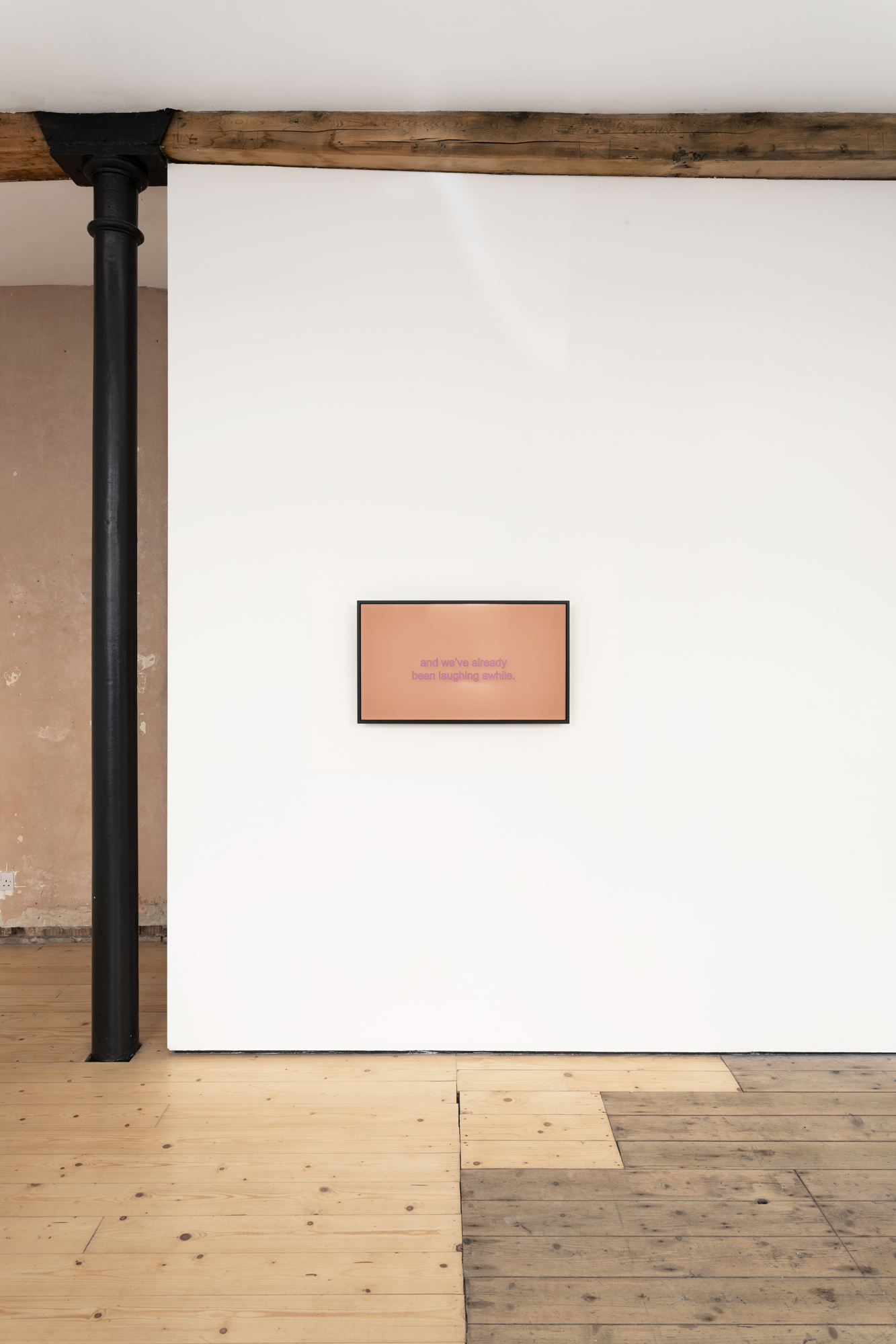 A screen placed on a white gallery wall, to the left of a dark grey pillar. The words 'and we've already been laughing awhile' appear on the screen.