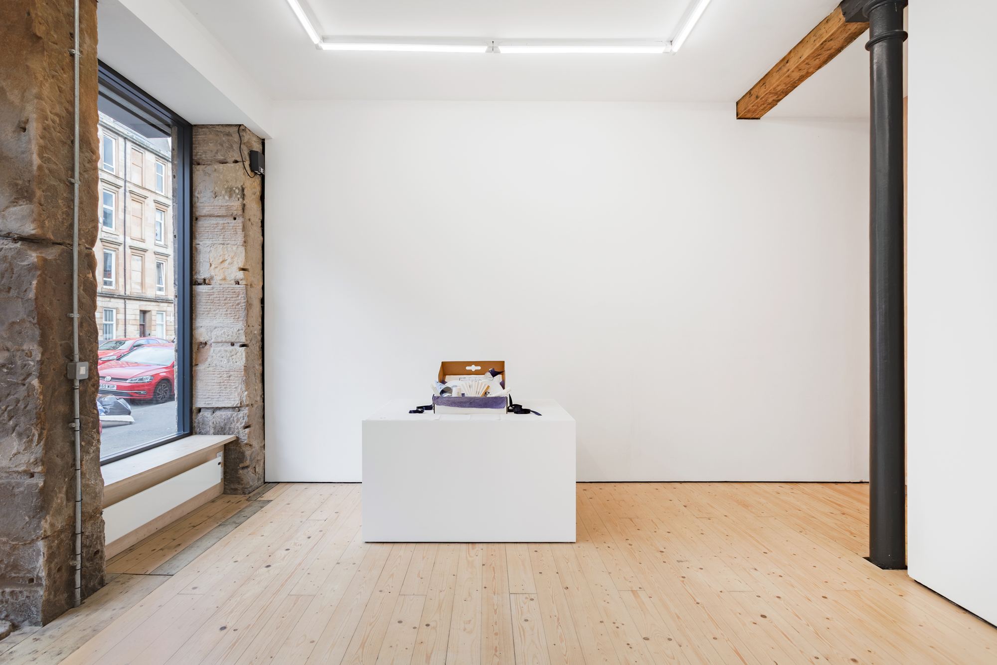 A large plinth and a screen placed in a white, window-lit gallery space. A cardboard box is placed on the plinth, a number of objects spilling out from this.