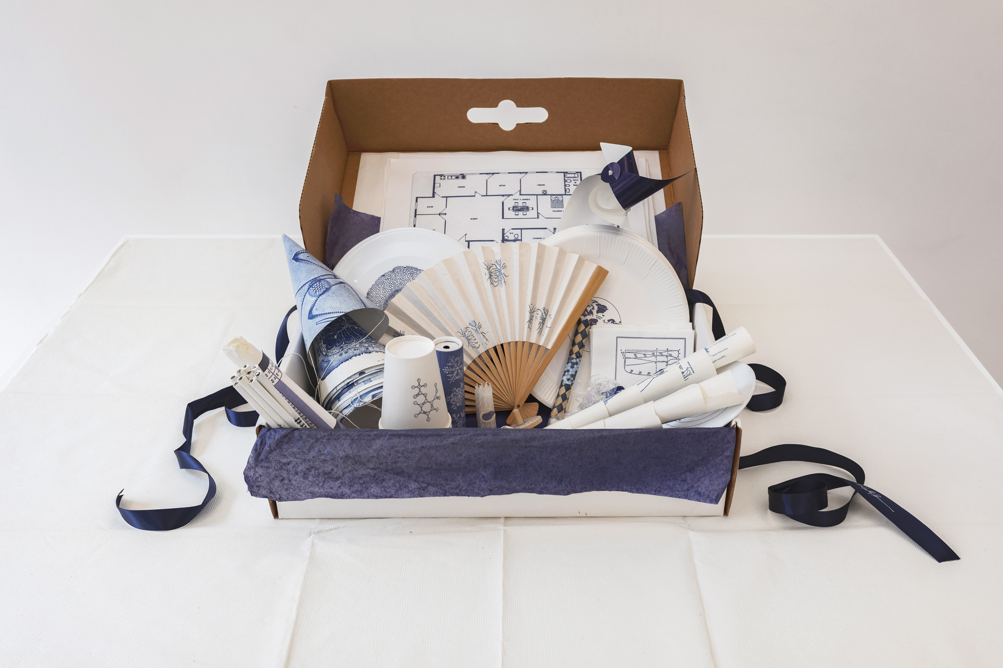 A white cardboard box inside which a variety of objects have been placed. These include party hats, candles, cups, pencils, a fan, and a pinwheel.