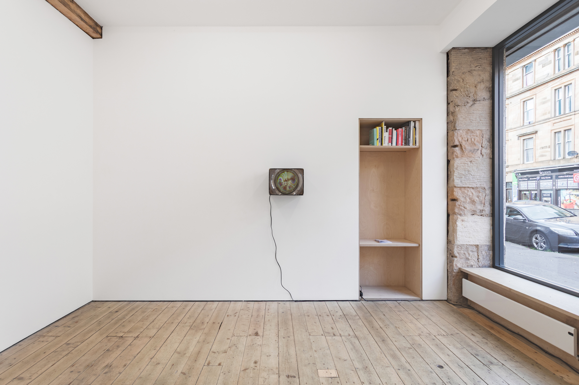 A box-like object hung on the wall of a white, window-lit gallery space, the front of which is a screen. The image that appears on the screen is heavily distorted. A cable trails from this object to the floor.