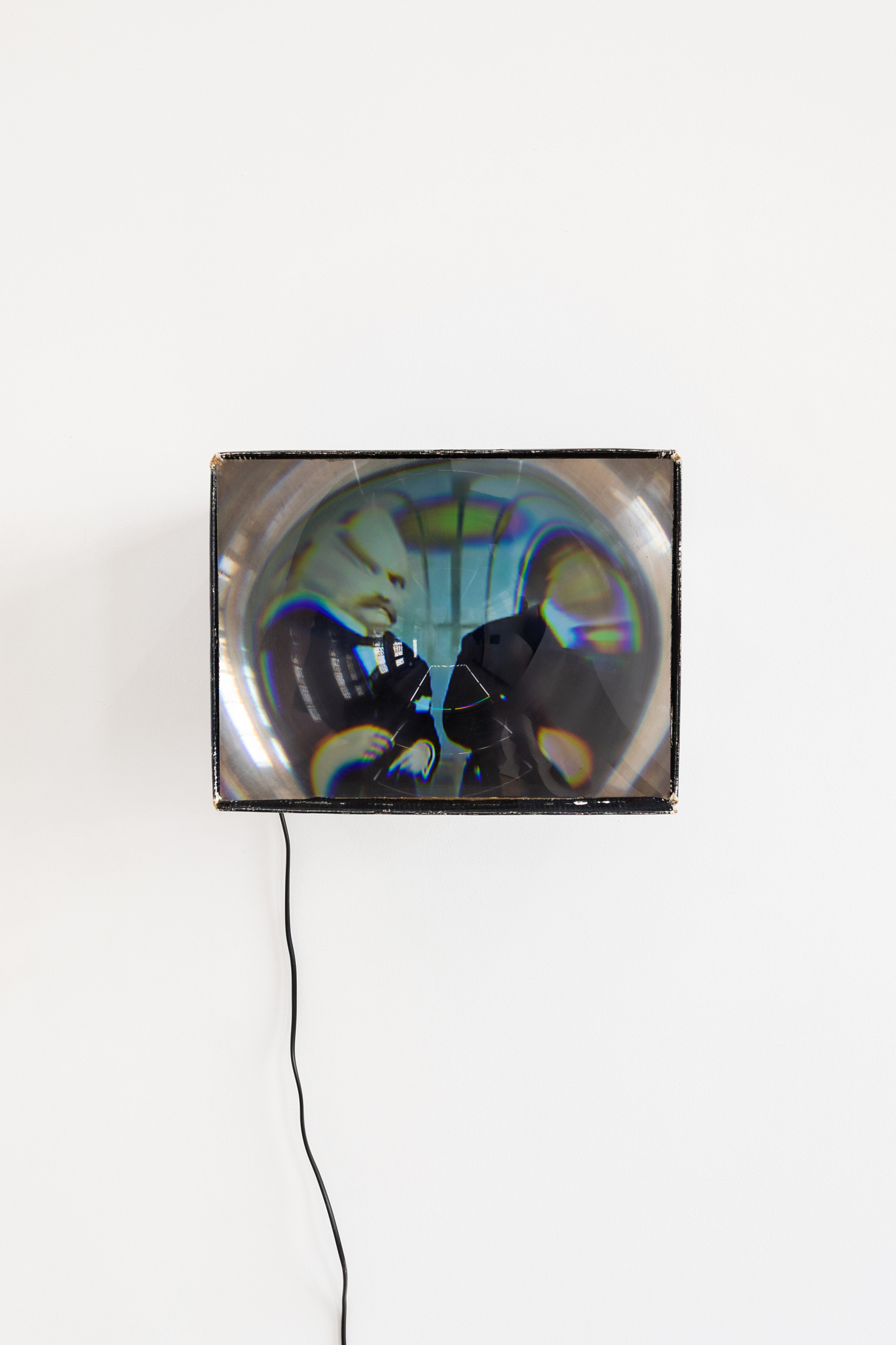 A frayed cardboard box hung on a white wall, with a plastic screen placed on its facing edge. The image that appears on the screen, of two men dressed in formal suits, is heavily distorted. A cable trails from this object to the floor.