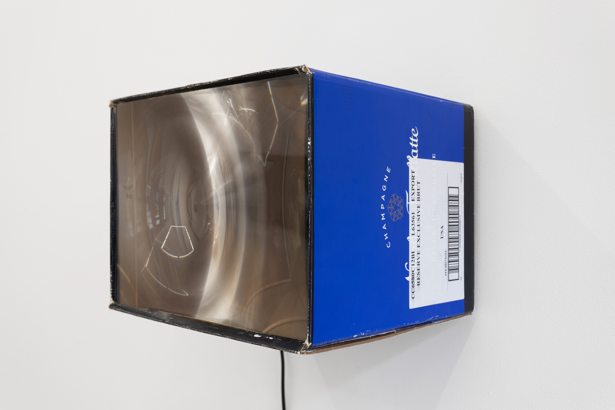 A frayed cardboard box hung on a white wall, with a plastic screen placed on its facing edge.The plastic screen displays a distorted reflection. Legible on the cardboard box are the words 'Champagne' and 'Reserve Exclusive Brut'.