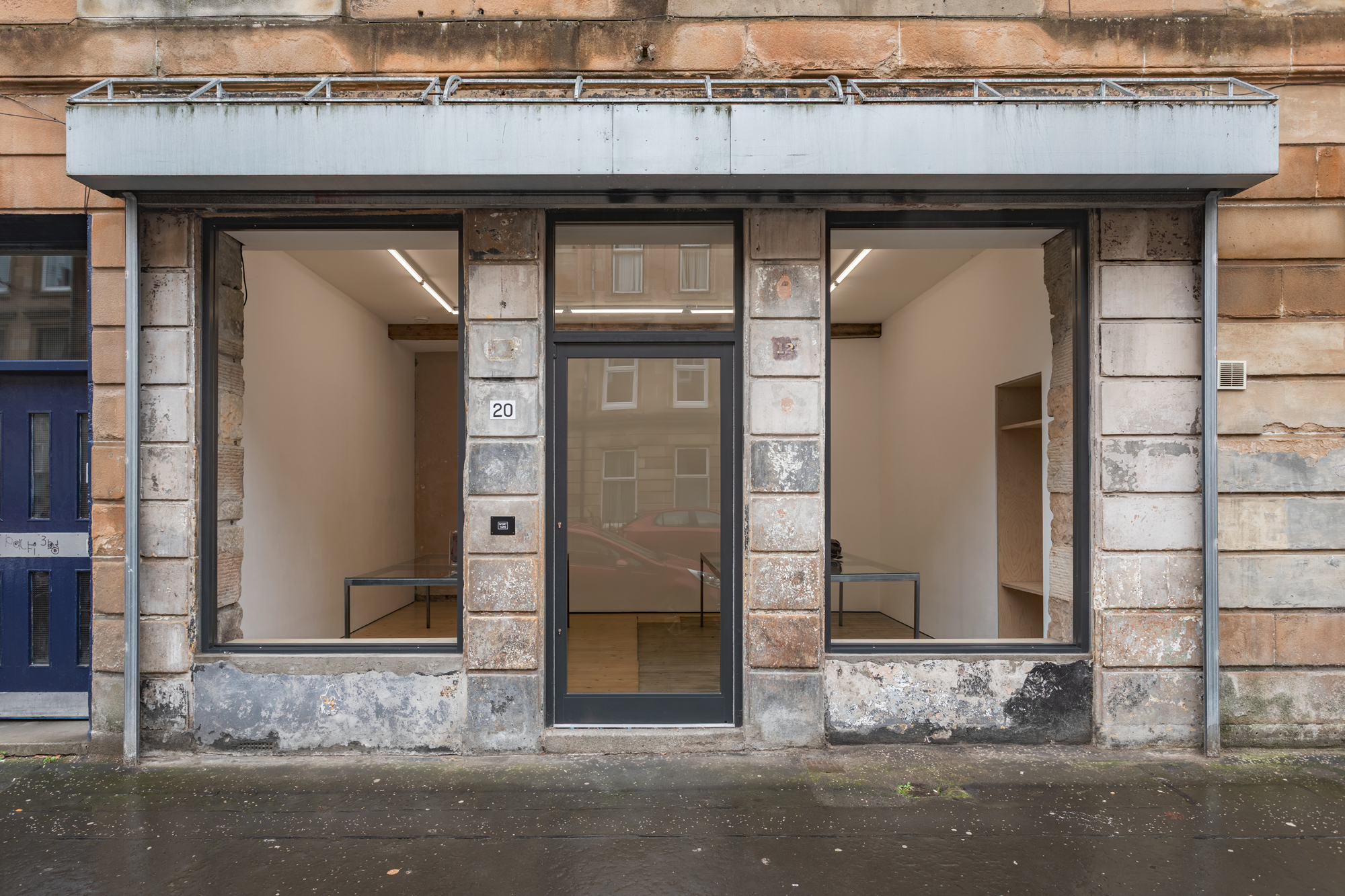 The facade of a gallery, from which the window panes have been removed. Two tables are partly visible through the openings.