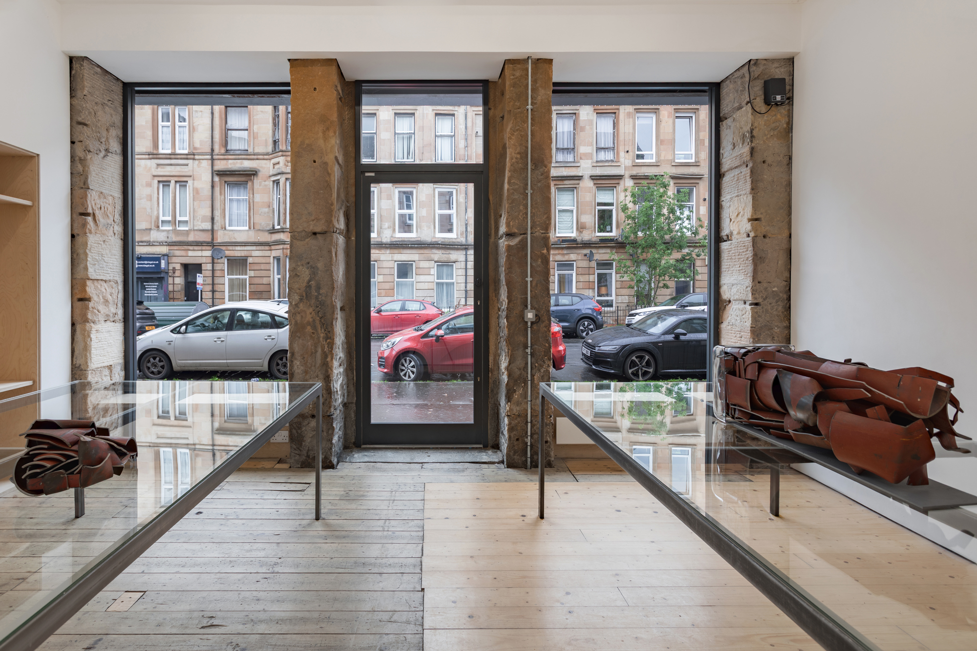 Two tables facing a a set of windows and a door. Sitting upon the tables are two red metal sculptures.