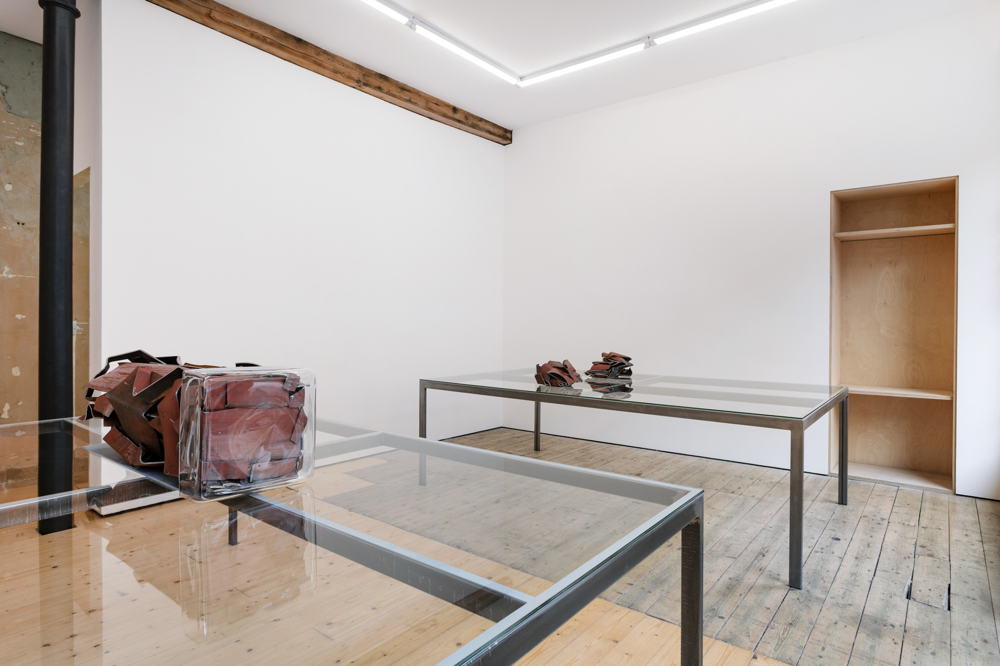 Two tables with glass surfaces in a gallery space, upon which three red metal sculptures have been placed.