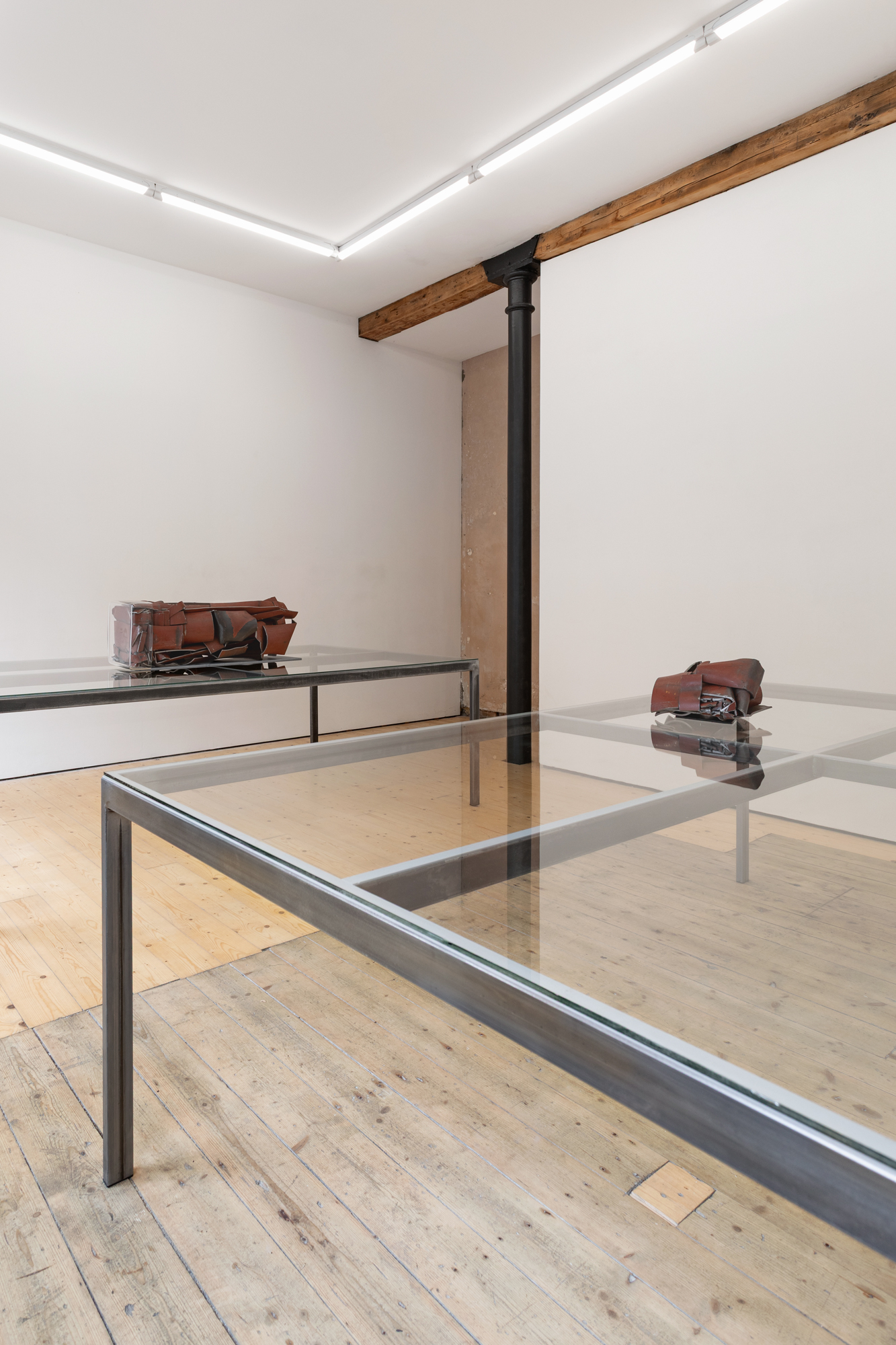 Two tables with glass surfaces in a gallery space, upon which two red metal sculptures have been placed.