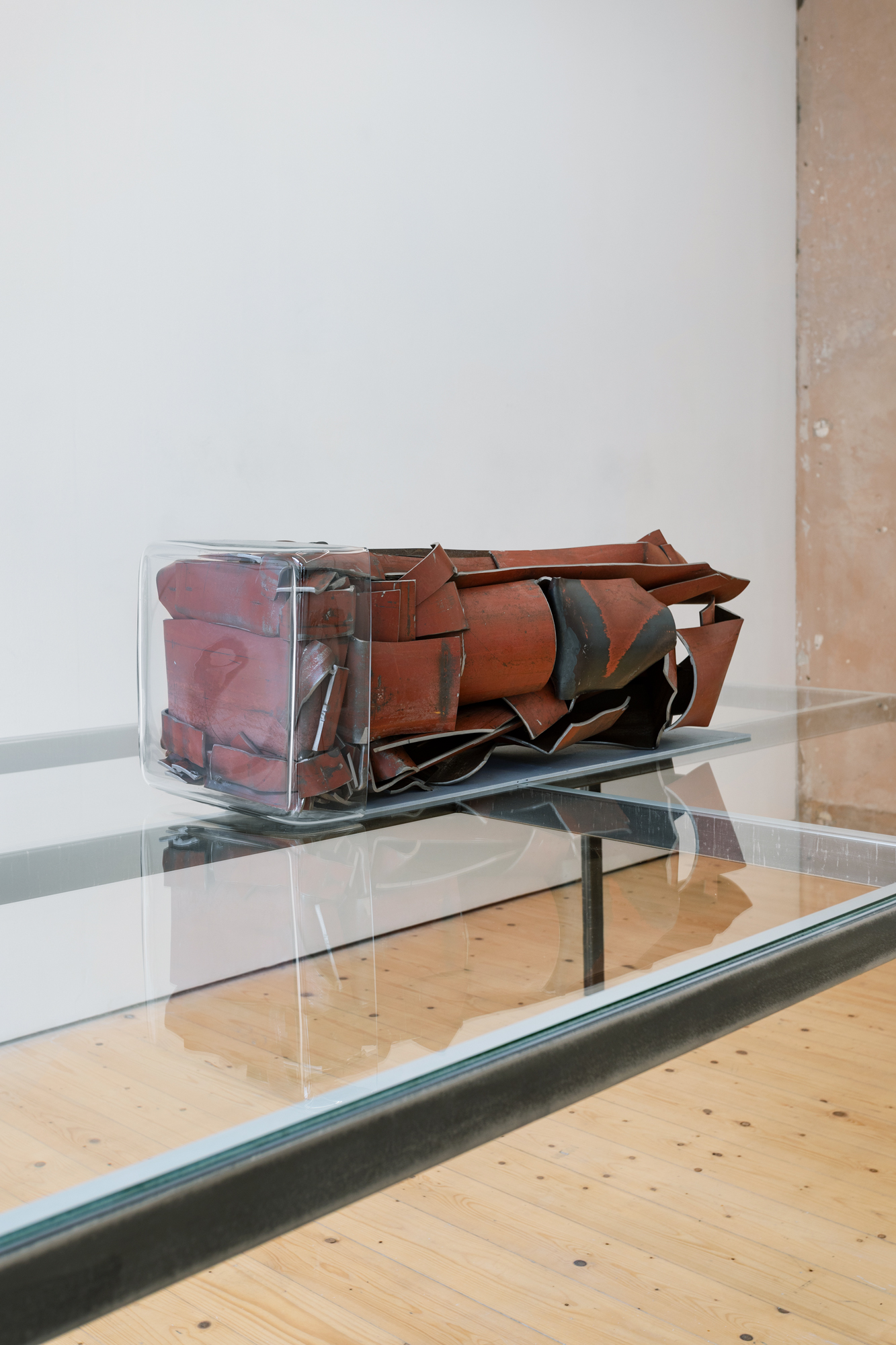 A sculpture made of red metal sitting on a glass surface. Another piece of glass is attached to the front of the sculpture.